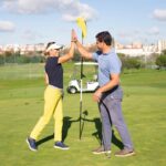 Two golfers high-five on the putting green beside a flagstick during a sunny training session.