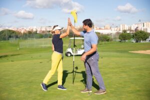 Two golfers high-five on the putting green beside a flagstick during a sunny training session.