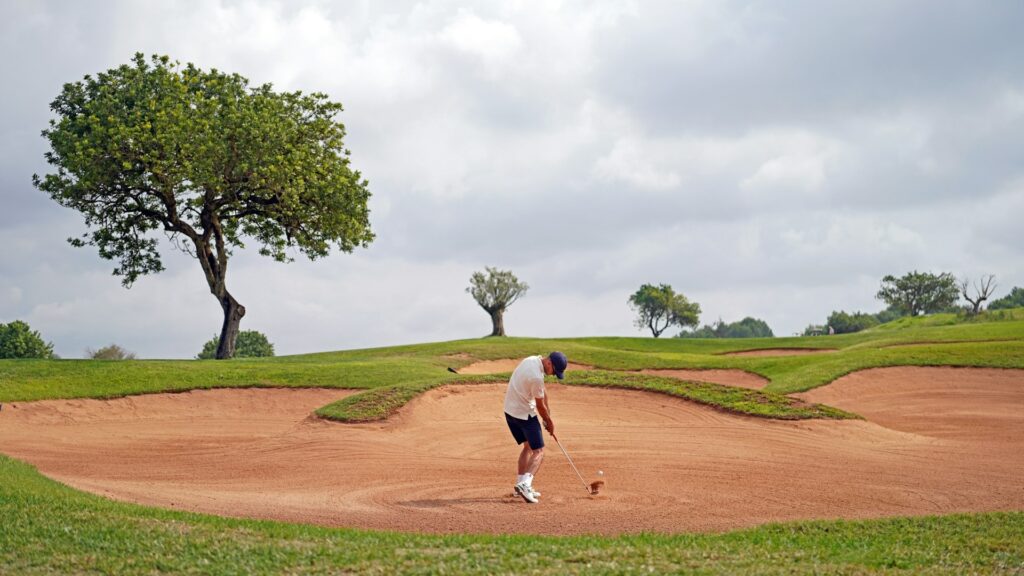 Golfer practicing a bunker shot from a sand trap on a cloudy day, showing golf training fitness for stability, technique, and lower-body control.