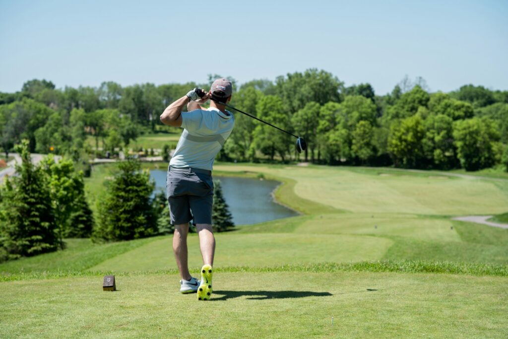 Golfer finishing a tee shot on a sunny course with a water hazard and fairway ahead, showing golf training fitness in action through balance and follow-through.