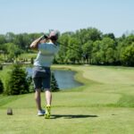 Golfer finishing a tee shot on a sunny course with a water hazard and fairway ahead, showing golf training fitness in action through balance and follow-through.