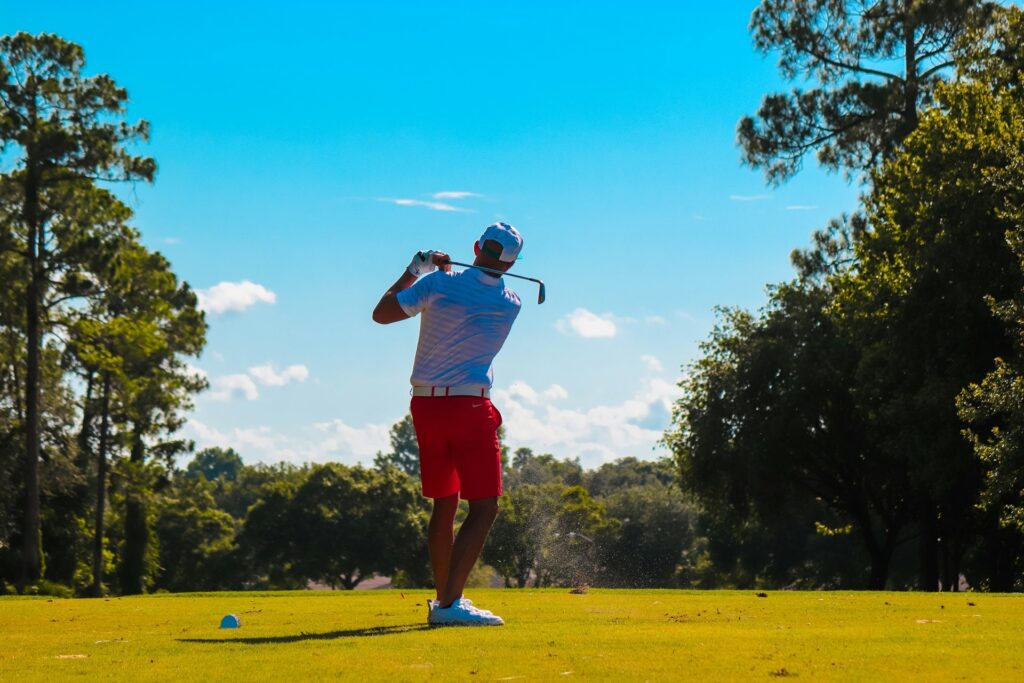 Golfer in red shorts swinging an iron under a bright blue sky on a tree-lined course, highlighting golf training fitness for power and control.