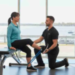 A physical therapist kneels beside an exam table and guides a seated patient through a resistance band leg test, showing how do physical therapists diagnose movement-related pain in a bright rehab clinic.