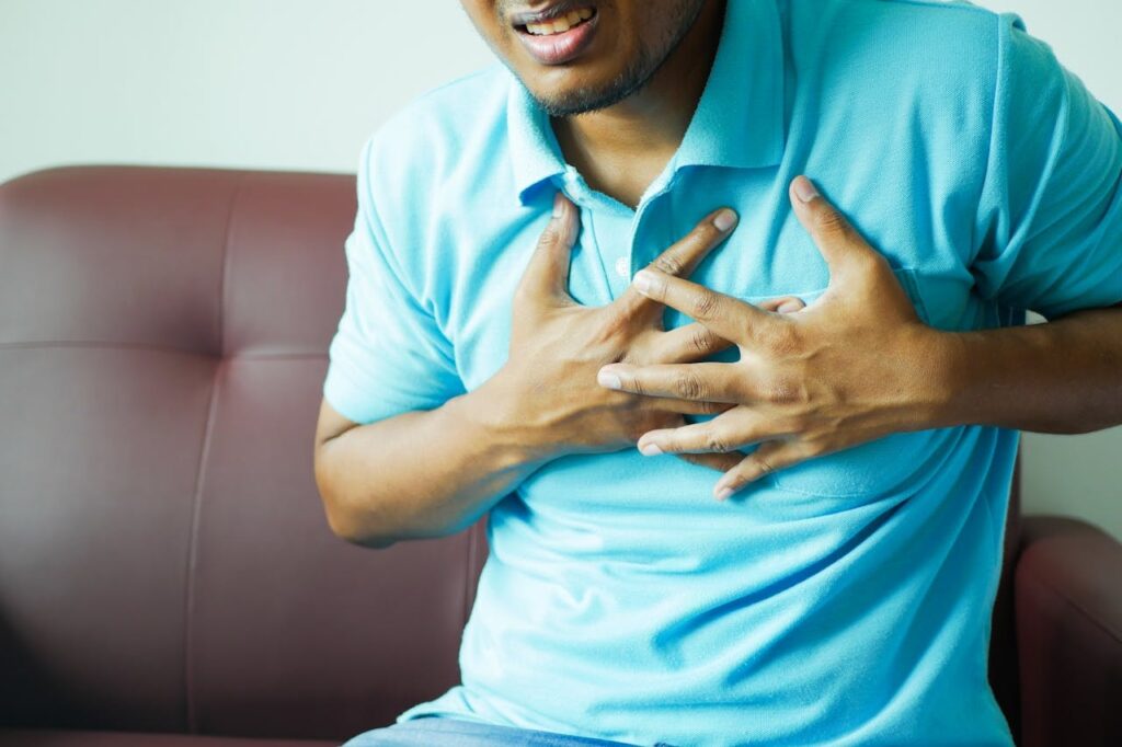 left chest pain numb arm as a man in a blue shirt clutches his chest while sitting on a couch