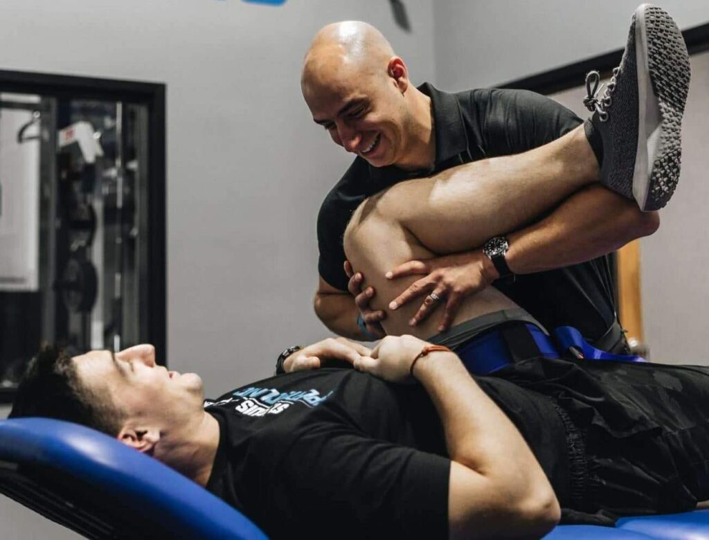 Professional sports therapist performing a hands-on hip and leg mobility stretch on an athlete lying on a treatment table in a gym-style clinic.