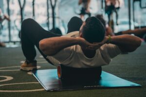 Spinal decompression workout in a gym, showing a person lying on a mat with a foam roller under the upper back, hands behind the head, using gentle pressure to relieve back tightness.