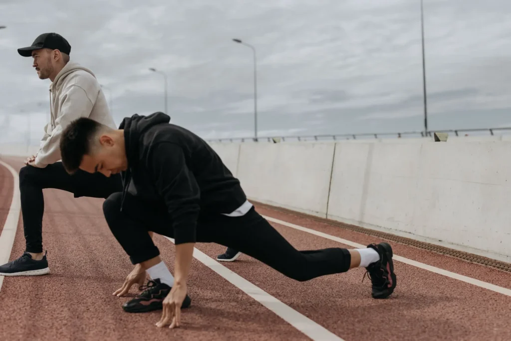 Stretching exercises for runners on a track, runner holding a deep lunge warm-up stretch while a training partner stands nearby