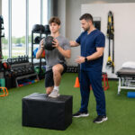 Athletic conditioning training in a physical therapy clinic, with a therapist coaching an athlete through a single-leg squat onto a box.