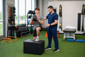 Athletic conditioning training in a physical therapy clinic, with a therapist coaching an athlete through a single-leg squat onto a box.